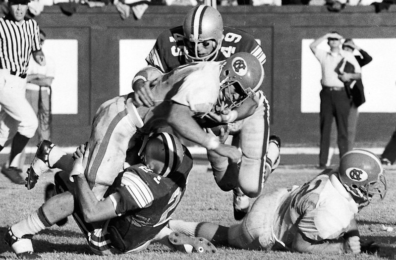 Football players during the 1973 Duke UNC match-up. Duke won it 27-10 after a tough loss in 1972. Pictured here: Duke's Weldon Baird (49) and David Hill (21) tackle Carolina's Mike Voight. Repository: Duke University Archives. Durham, North Carolina, USA. library.duke.edu/uarchives