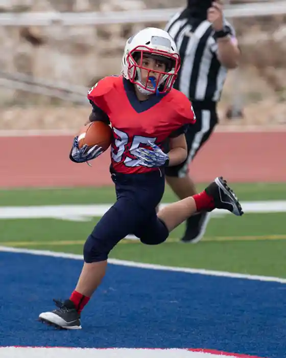 Youth player running for a touch. Wearing a red jersey with blue pants. Referee in the background.